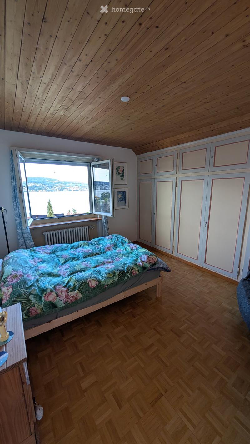 Bright bedroom with wooden herringbone flooring, slanted wooden ceiling, large window offering a scenic view, bed with floral duvet, wardrobe, radiator, and small fan on nightstand.
