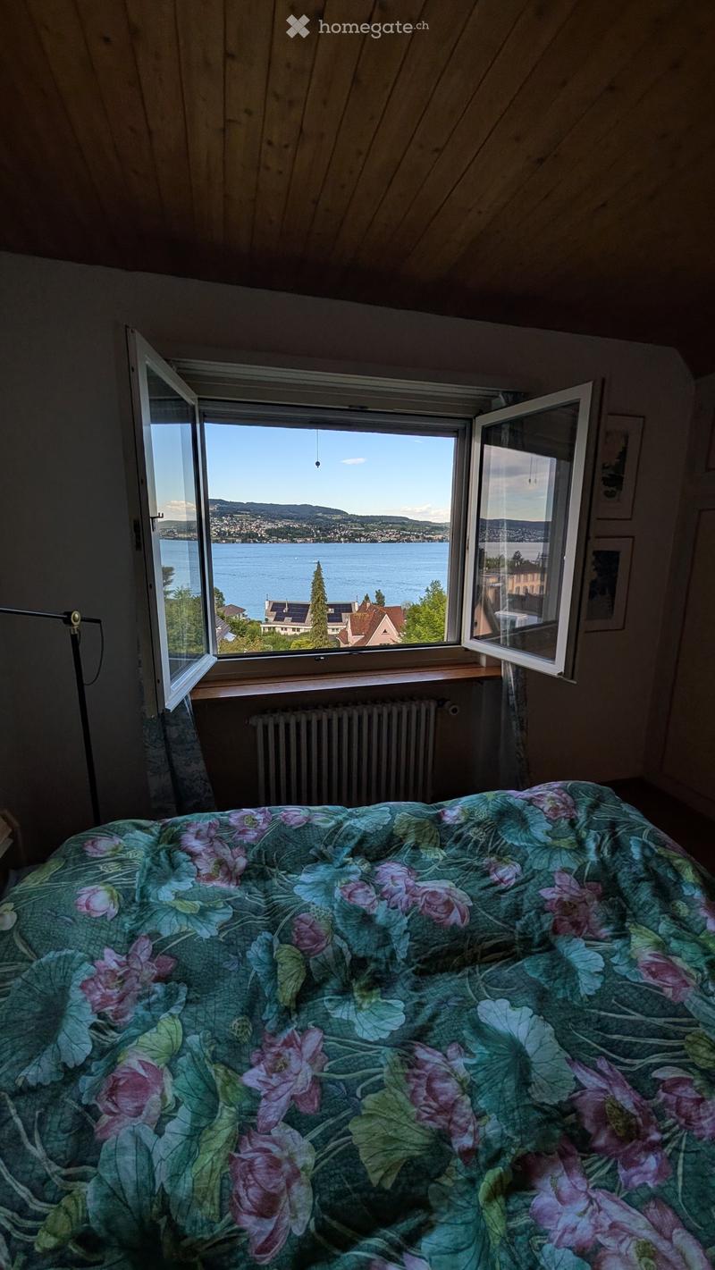 Bedroom with a large window offering a scenic view of a lake and surrounding landscape. The room features a bed with a floral-patterned duvet, a radiator below the window, and a floor lamp near the window.