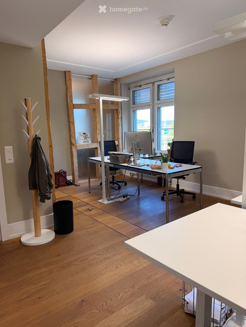 Modern office space with wooden flooring, large windows, a desk with a computer monitor, office chairs, a coat rack, and a small plant.