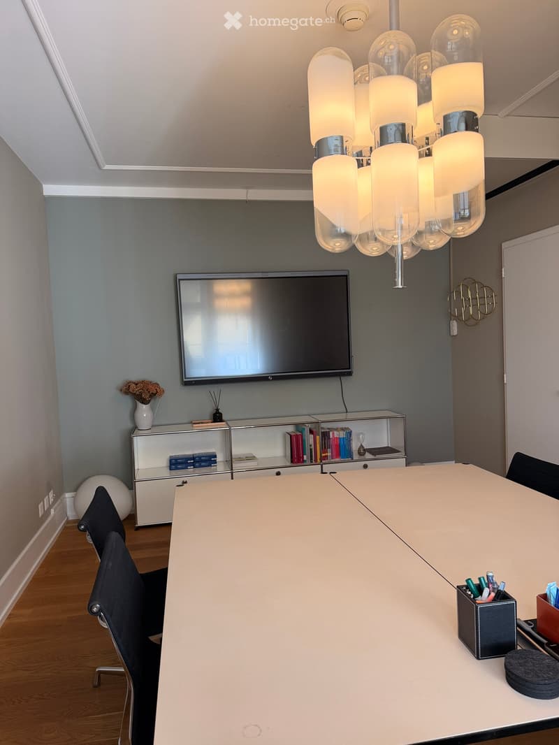 Modern conference room with a large white table, black chairs, a mounted TV screen, a bookshelf with books and decorative items, and a pen holder on the table.