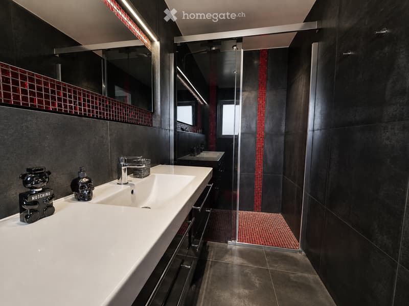 Modern bathroom with double sink, sleek black tiles, red mosaic accents, and a glass-enclosed shower area.