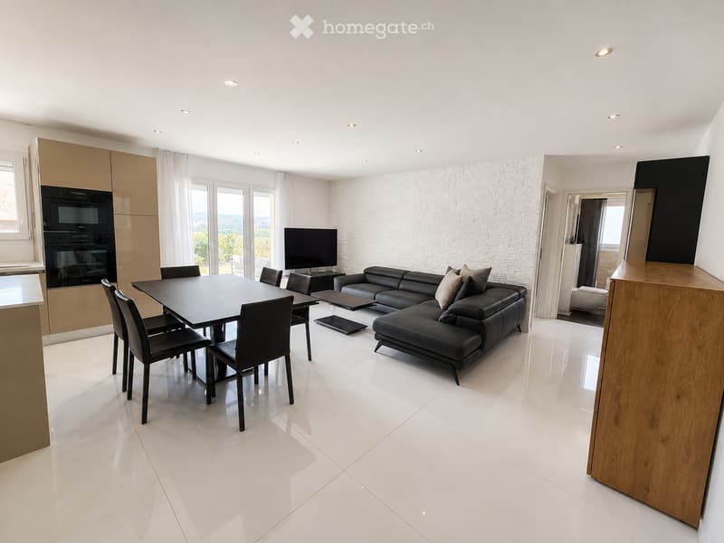 Modern living and dining area with white tiled flooring, a black leather sofa, a dining table with chairs, and a television. The space features large windows with white curtains and a view of greenery outside.