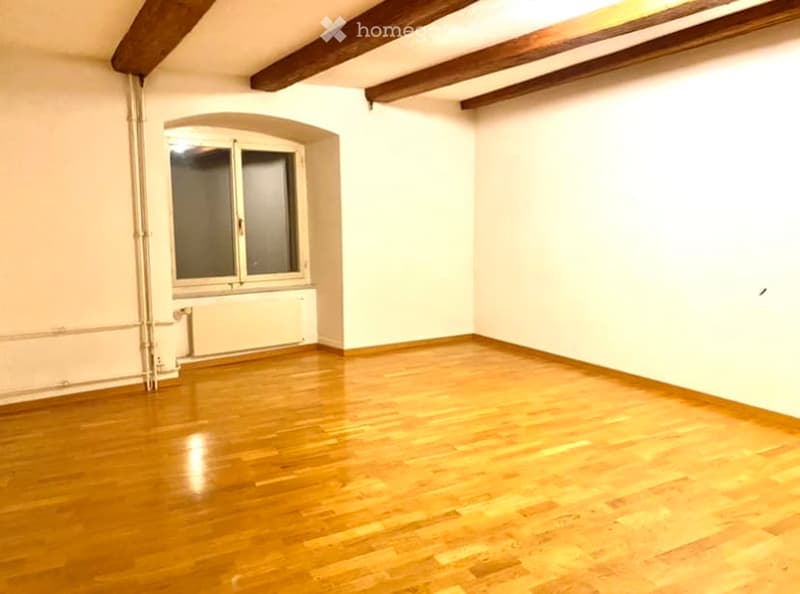 Empty room with wooden flooring, white walls, and exposed wooden beams on the ceiling. Features a single window with double panes and a radiator below it.