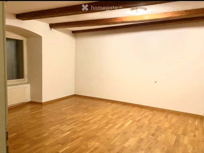 Empty room with wooden flooring, white walls, and exposed wooden beams on the ceiling. A single window with a radiator below it is visible on one wall.