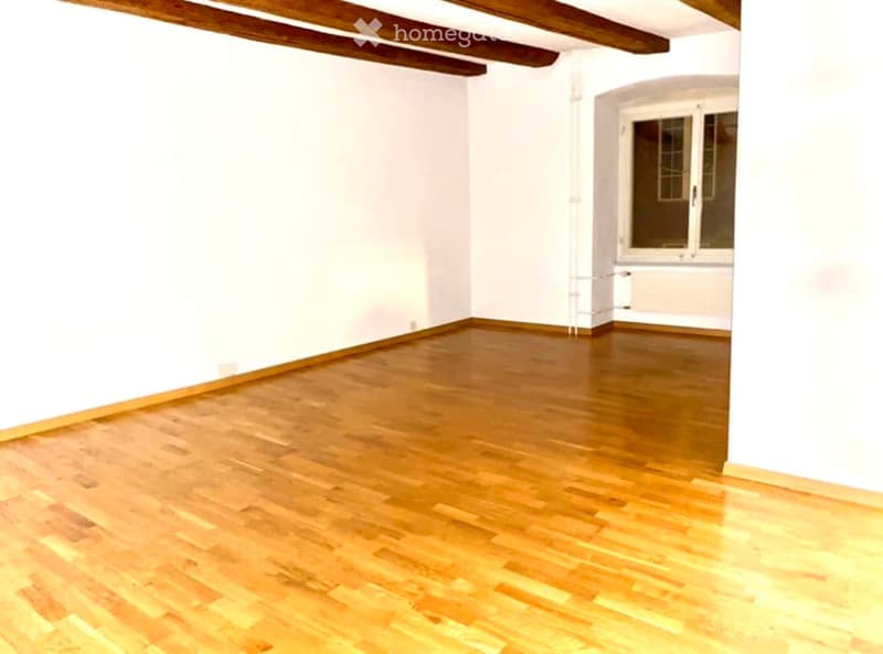 Empty room with wooden flooring and white walls, featuring exposed wooden beams on the ceiling and a single window with a white frame.