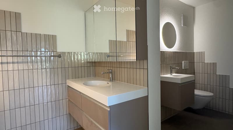 Modern bathroom with two sinks, one with a large mirror and the other with a built-in cabinet, both featuring sleek, contemporary design and neutral tilework.