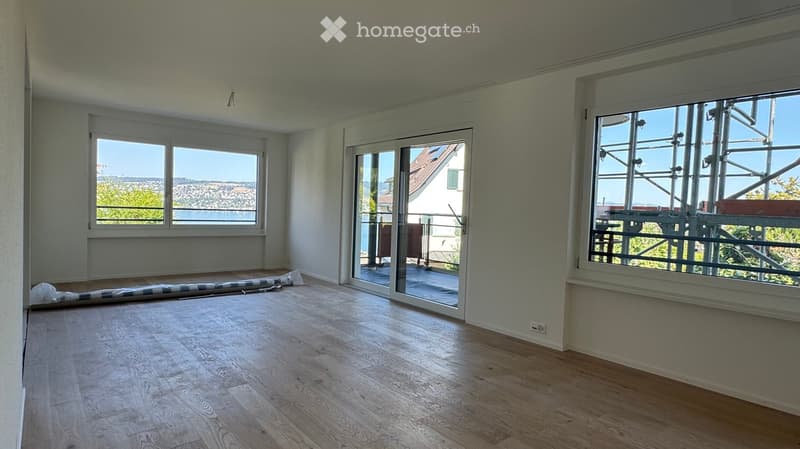 Empty room with wooden flooring, large windows, and a sliding glass door leading to a balcony. The room has white walls and a view of the lake and surrounding landscape.