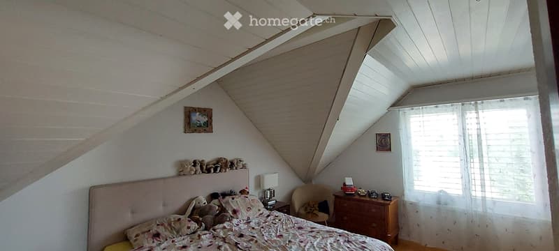 Cozy attic bedroom with slanted ceilings, a bed with a floral comforter, a nightstand with a lamp, a small chair, and a window with blinds.