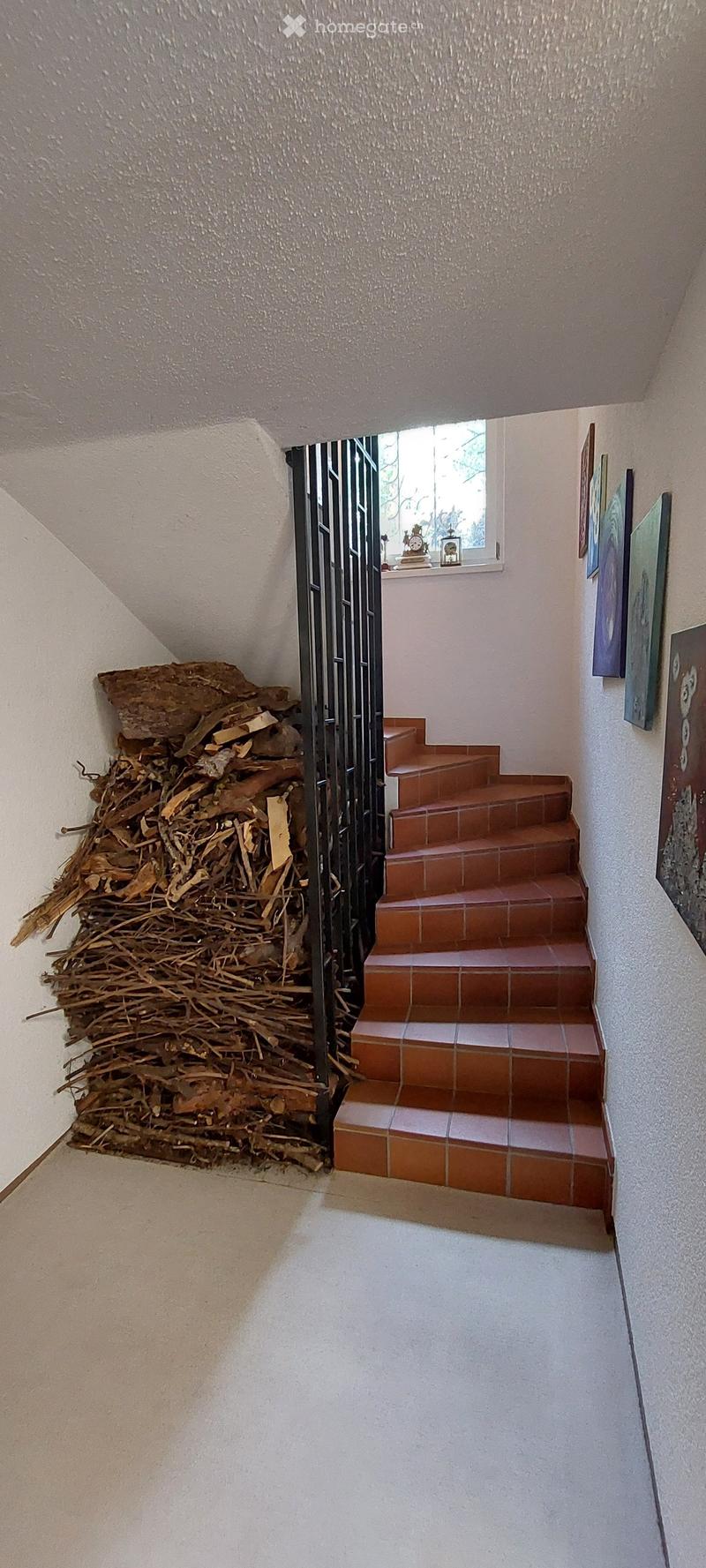 A narrow hallway with a staircase leading upwards, featuring a metal gate, a pile of firewood on the left, and a small window at the top with a few decorative items on the sill.
