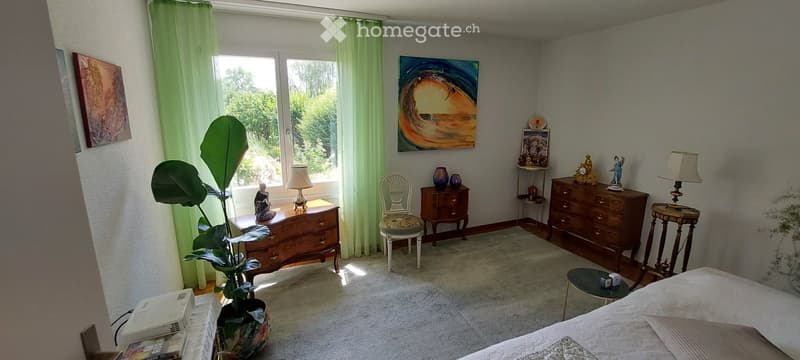 Bright bedroom with large windows, white walls, and wooden furniture. Features a bed, nightstand, lamp, chair, small table, plant, and artwork.