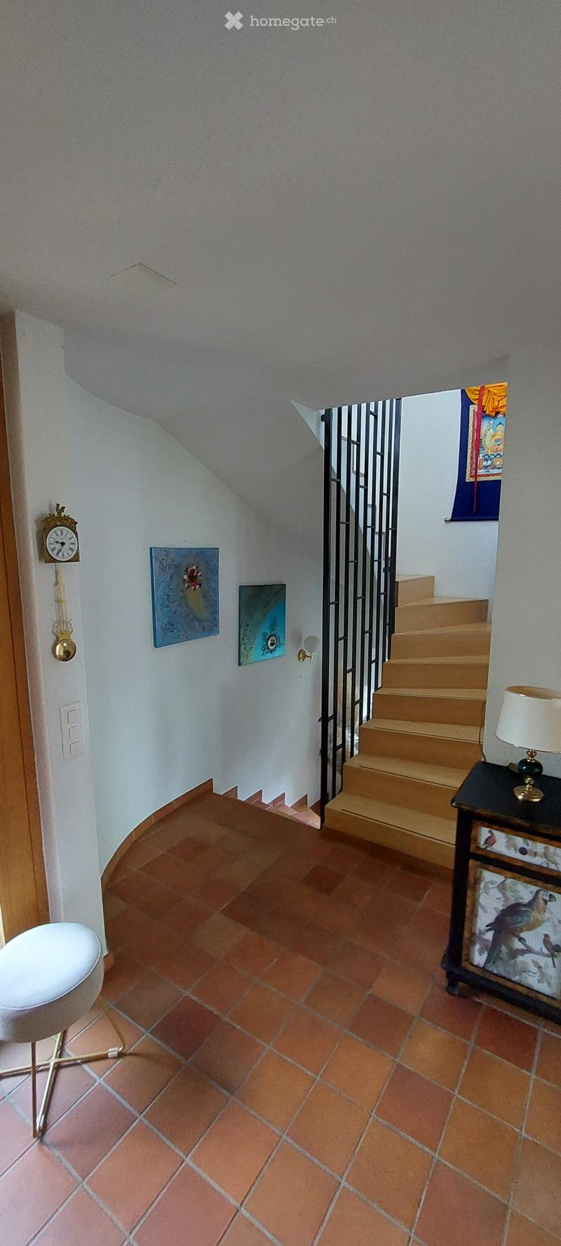Hallway with terracotta tiled floor, white walls, staircase leading upwards, small table with lamp, wall-mounted clock, and artwork on walls.