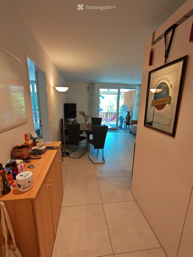 A well-lit hallway leading to a dining area with a table, chairs, and a laptop. A framed picture hangs on the wall, and a small cabinet holds various items, including bottles and a mirror.