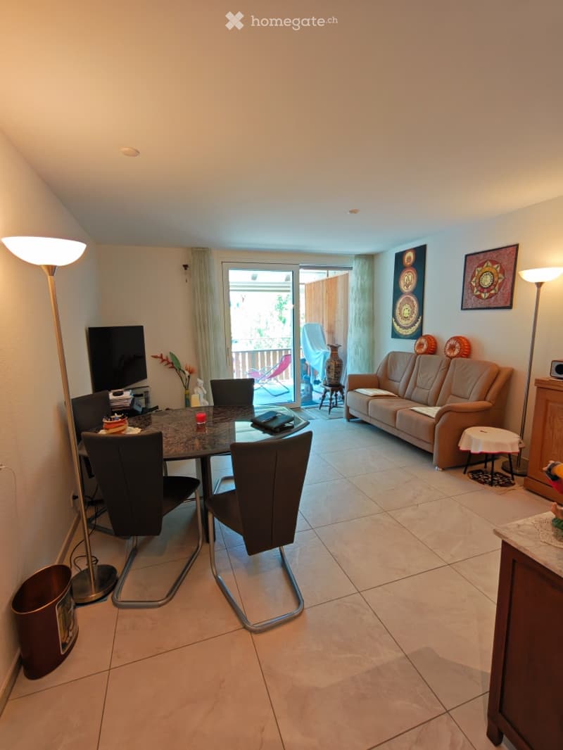 Bright living room with tiled floor, large sliding glass door leading to a balcony, beige sofa, coffee table, dining table with chairs, floor lamp, and decorative items.