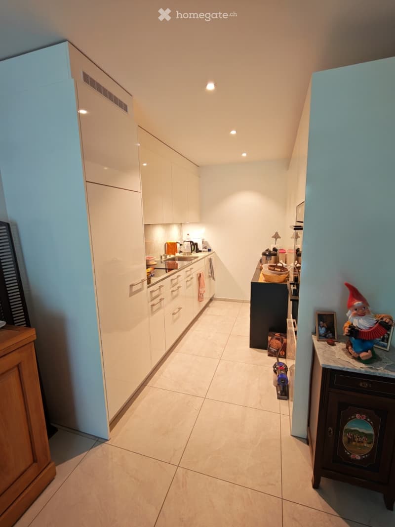Modern kitchen with white cabinets, a central island, and various appliances including a coffee maker and oven. The floor is tiled, and there is a small table with a chair and a decorative gnome figurine on a sideboard.