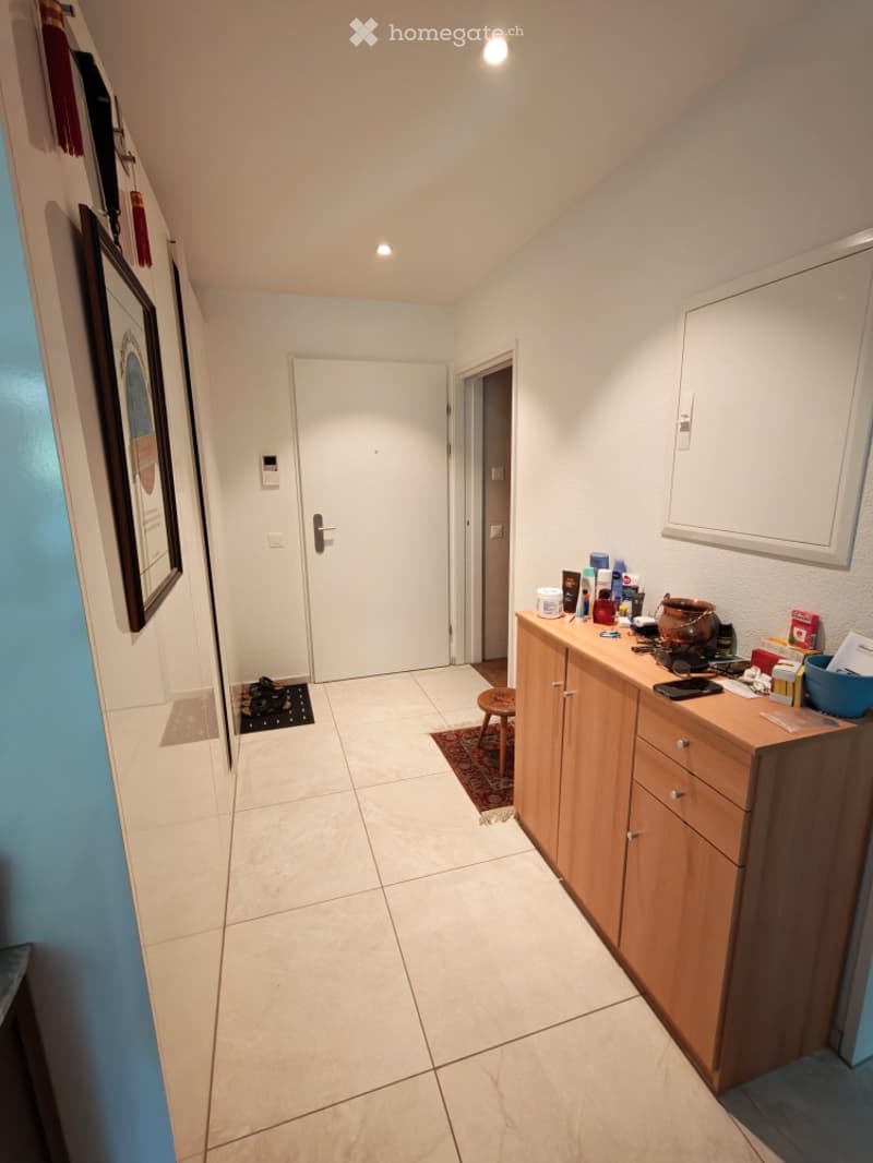 A narrow hallway with tiled flooring, white walls, and a wooden cabinet on the right side. A door leads to another room, and a small rug is placed near the entrance.
