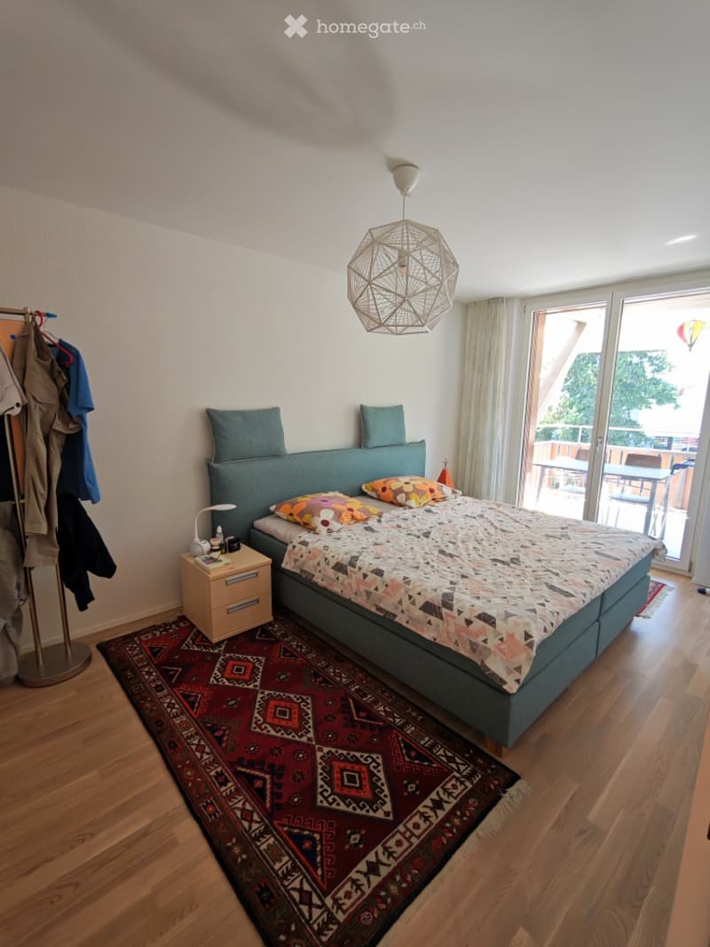 Modern bedroom with a large bed, colorful bedding, nightstand, lamp, and a patterned rug. A sliding glass door leads to a balcony.