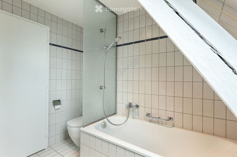 Modern bathroom with a bathtub, toilet, and glass-enclosed shower. The walls are tiled with white tiles and blue accents, and the floor is also tiled.