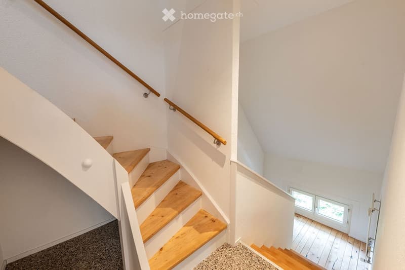Wooden staircase with white walls and a wooden handrail, leading to a room with wooden flooring and large windows.