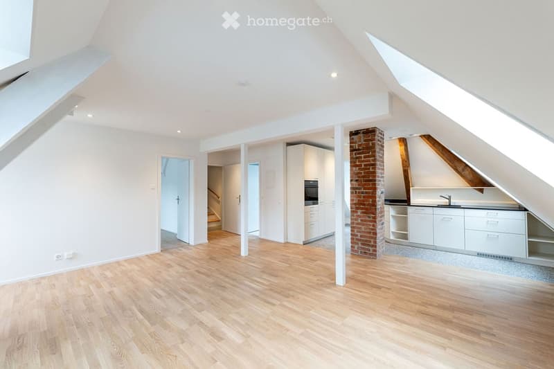 Bright, open attic space with wooden flooring, white walls, exposed brick pillar, and built-in kitchen cabinets. The room features large skylights and a staircase leading to another area.