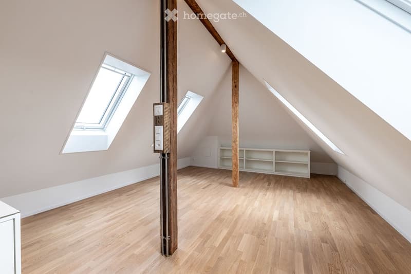 Empty attic room with wooden flooring, exposed wooden beams, and built-in white shelving unit. Skylight windows provide natural light.