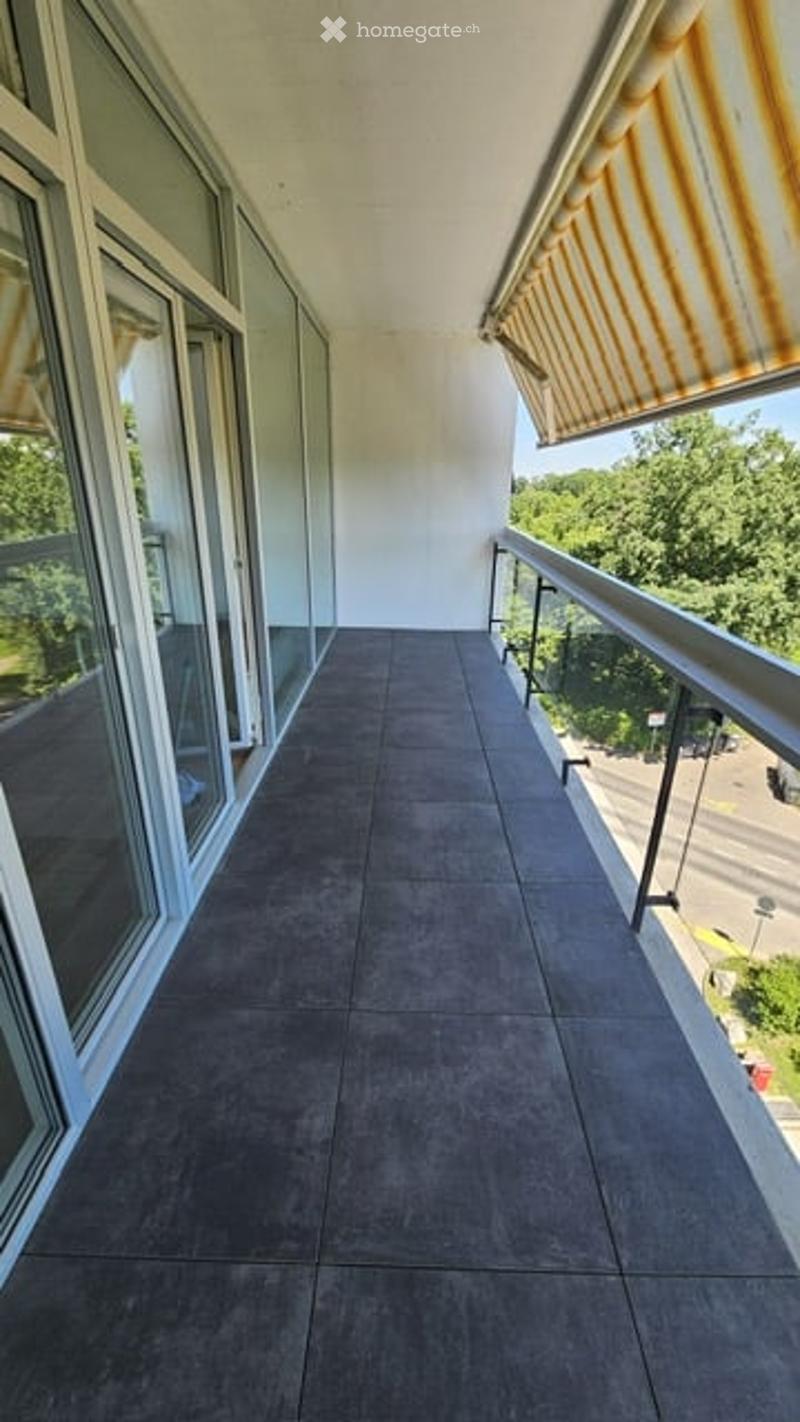 Balcony with grey tiled floor, white walls, large windows, metal railing, and striped awning. Offers a view of greenery and street.
