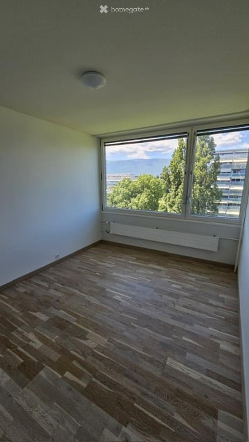 Empty room with wooden flooring, large window offering a view of trees and buildings, white walls, and a radiator under the window.