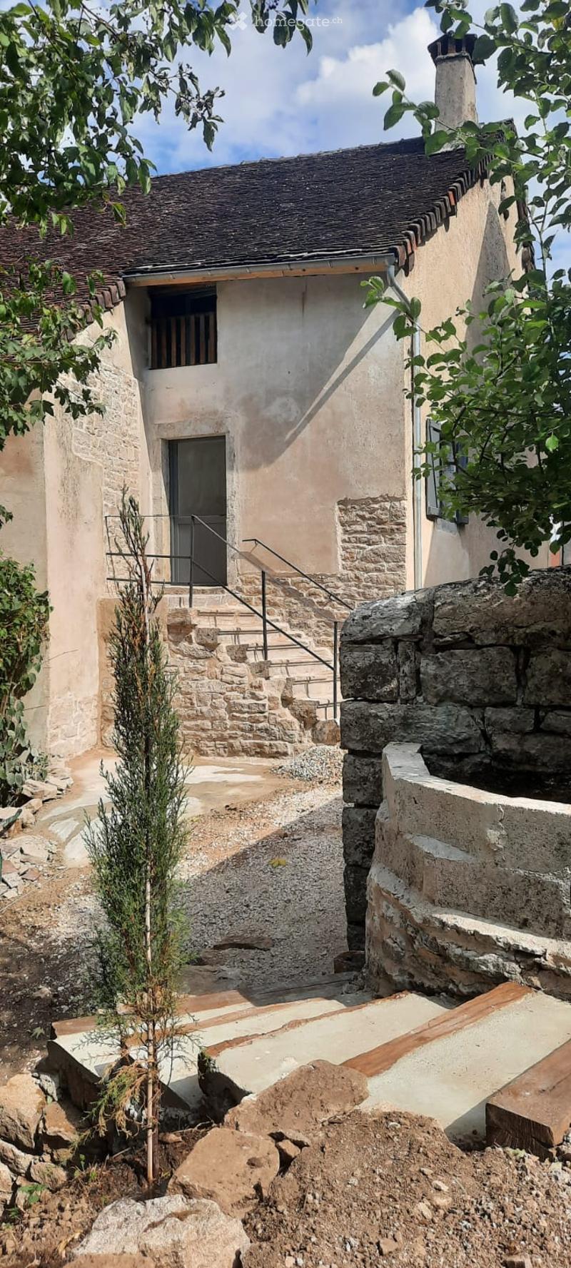 A rustic stone house with a tiled roof, featuring a small staircase leading to an entrance door, surrounded by stone steps and greenery.