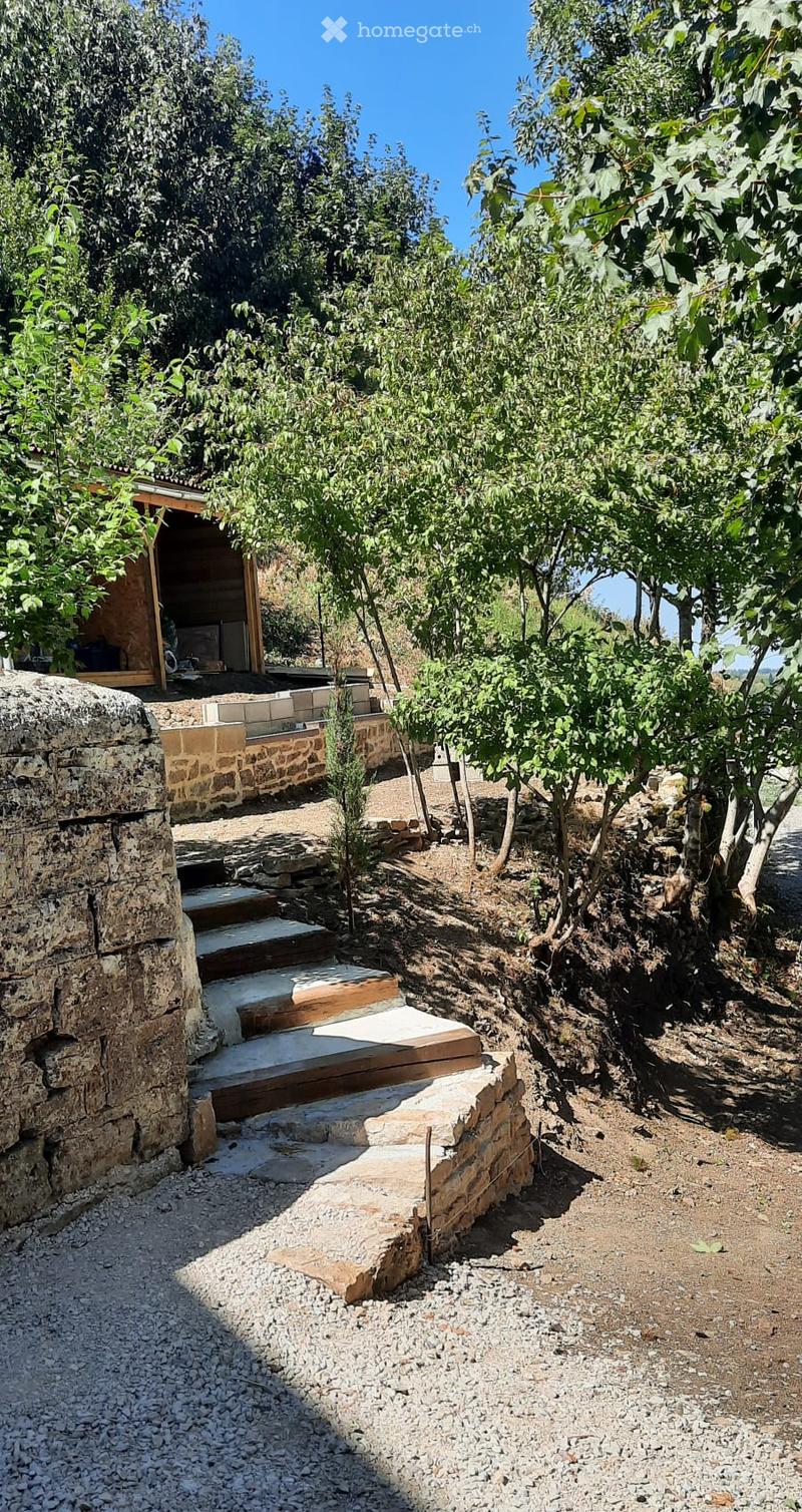 A rustic outdoor area with stone steps, gravel pathways, and a wooden platform leading to a covered structure surrounded by trees and greenery.