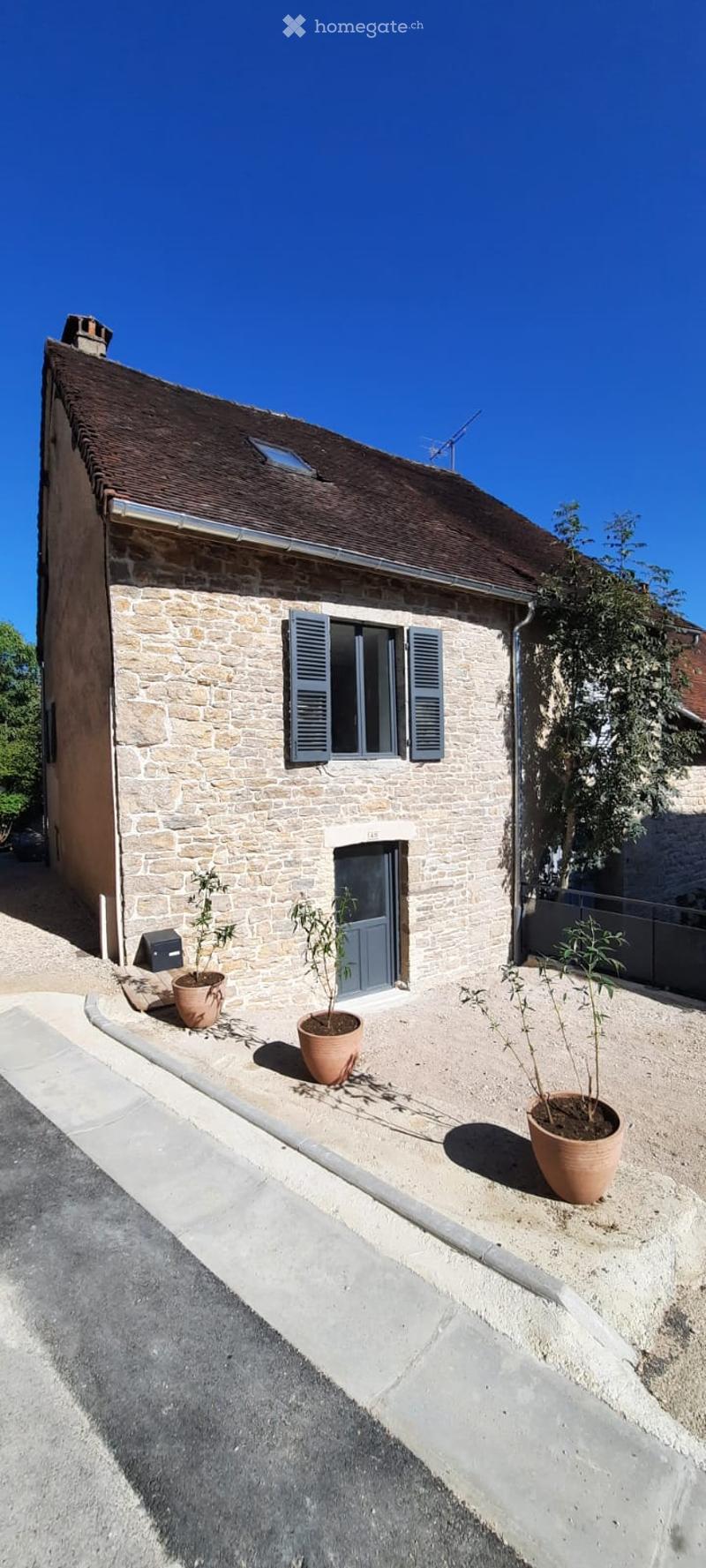 A stone house with a tiled roof, blue shutters, and a small garden area with potted plants.