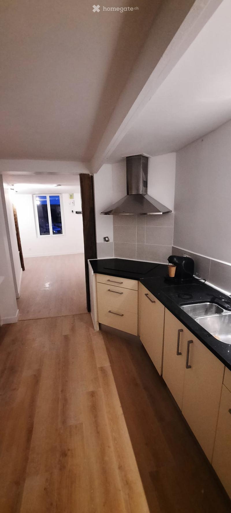 Modern kitchen with light wood cabinets, black countertops, stainless steel sink, and a range hood. The floor is wooden, and a small window is visible in the background.