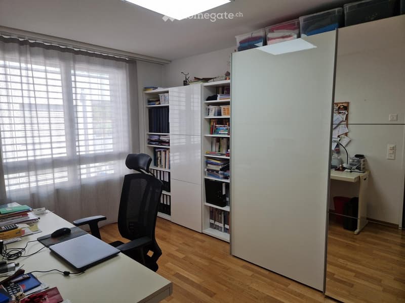 Office space with wooden flooring, large window with blinds, white bookshelf, desk with laptop, chair, and various office supplies.