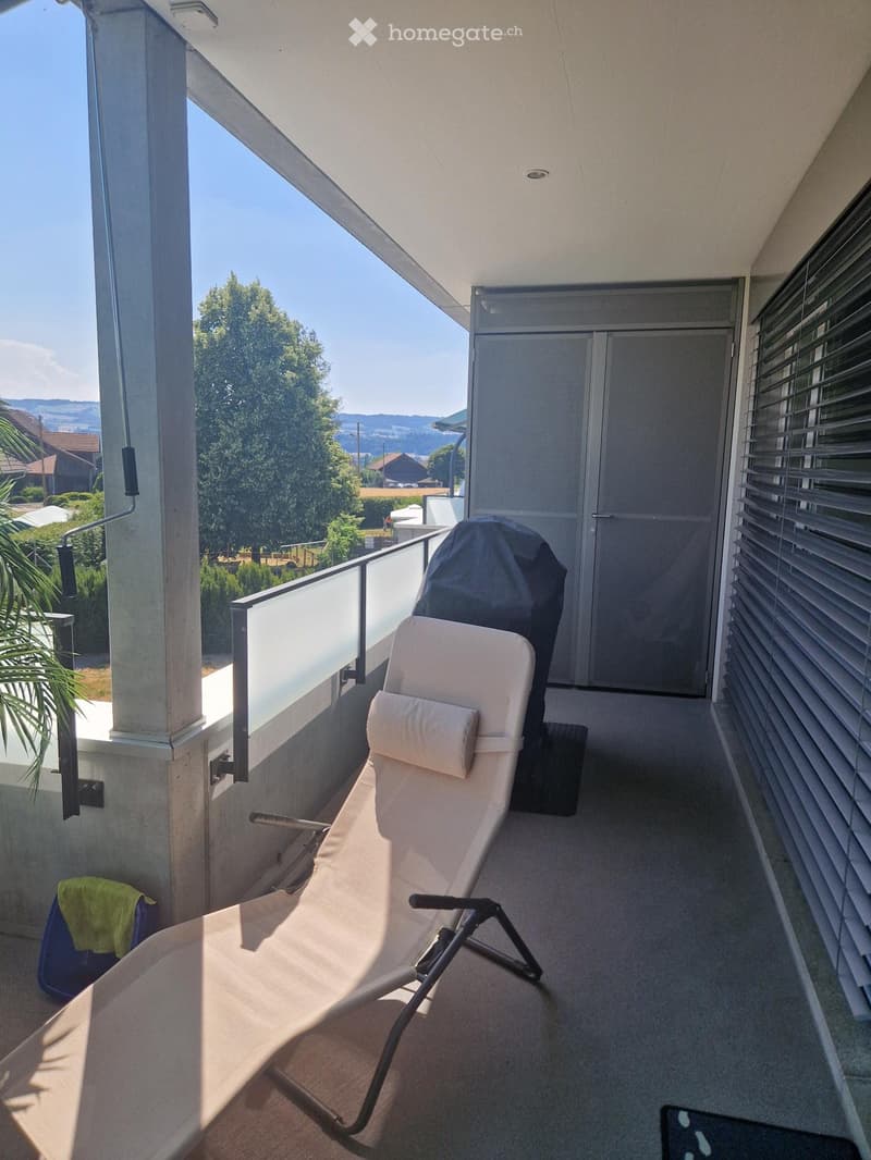 Balcony with a sun lounger, cushion, and covered grill, overlooking a scenic landscape with trees and hills.