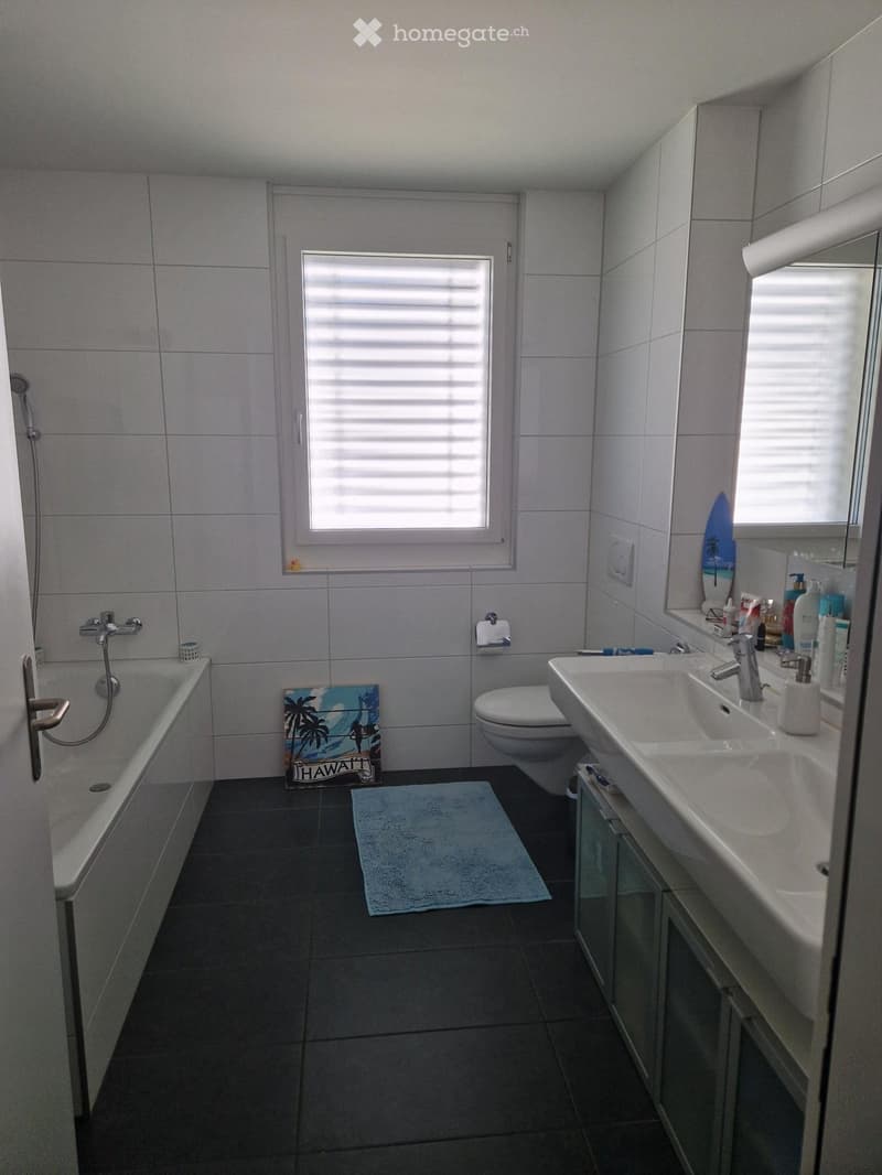 Modern bathroom with a bathtub, toilet, sink, mirror, and various toiletries. Features a window with blinds, a towel on the floor, and a decorative sign on the wall.