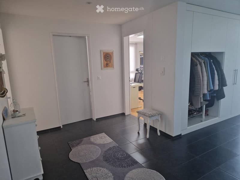 The image shows a well-lit hallway with a dark tiled floor, light-colored walls, and a built-in wardrobe with hanging clothes. A small round rug with a decorative pattern is placed on the floor, and a small white stool with a patterned seat is positioned