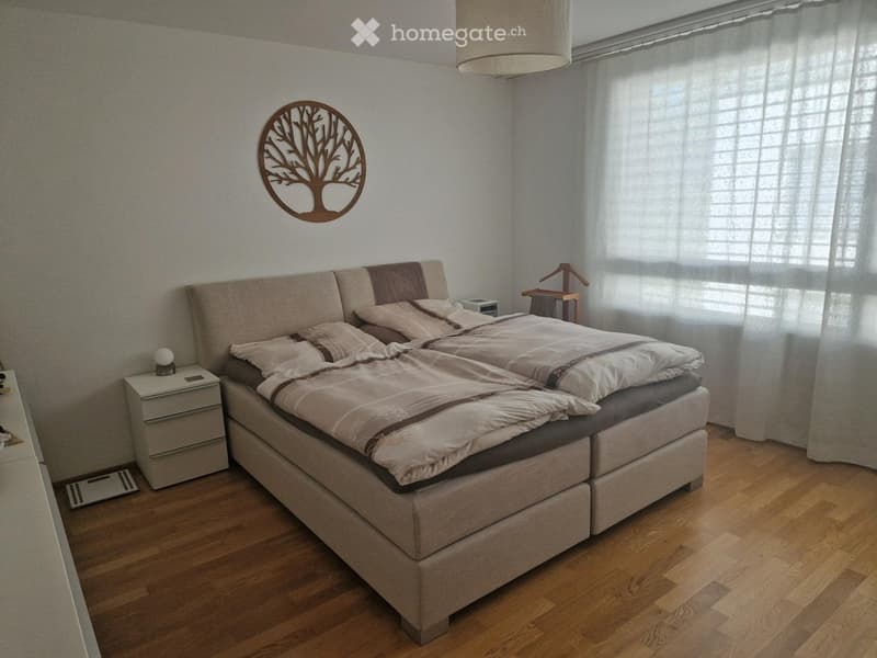 Modern bedroom with a large bed, nightstand, lamp, and a decorative wall mirror. Wooden flooring and large window with blinds.