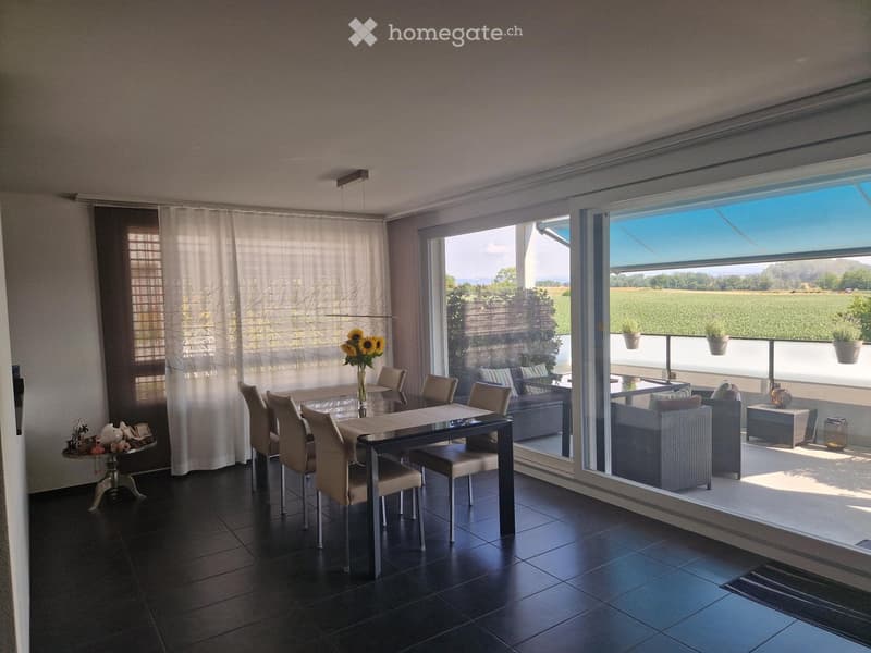 Bright dining area with a glass table, six chairs, and a view of a balcony with outdoor seating. The room features large windows, dark tiled flooring, and a decorative vase with sunflowers on the table.