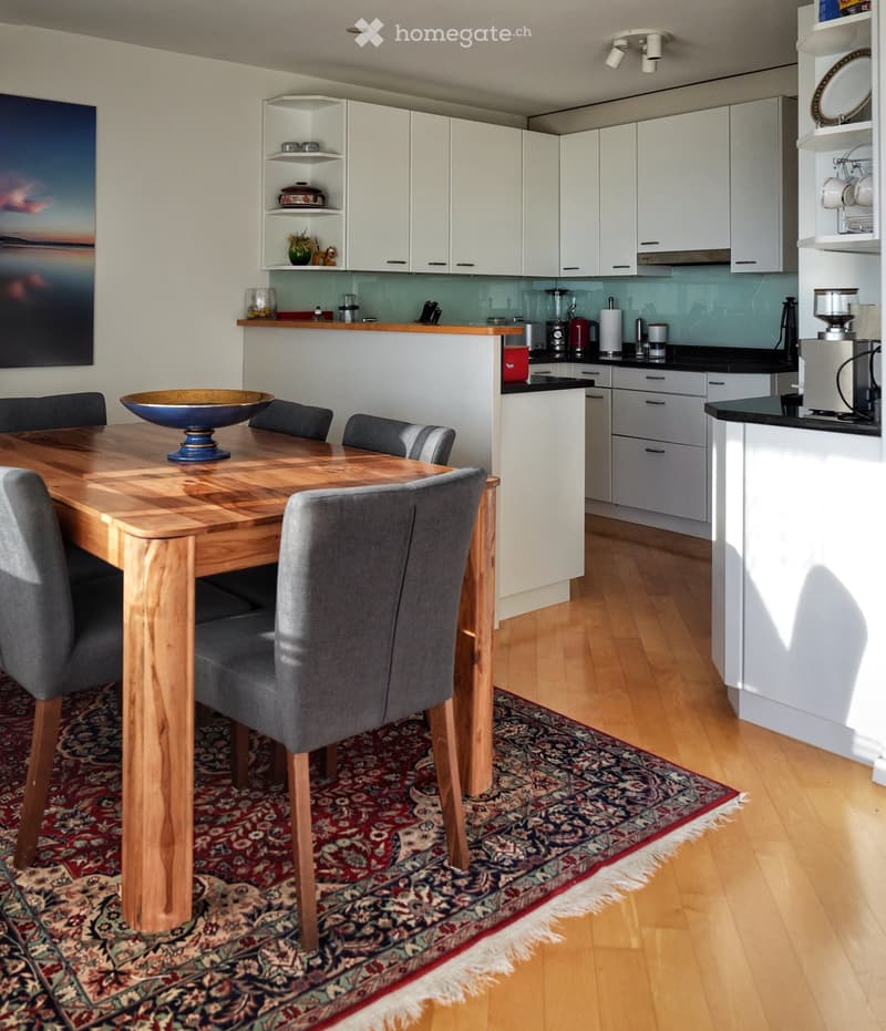 Modern kitchen with white cabinets, wooden dining table, grey chairs, and various appliances including a blender and coffee maker. A colorful rug is placed under the dining area.