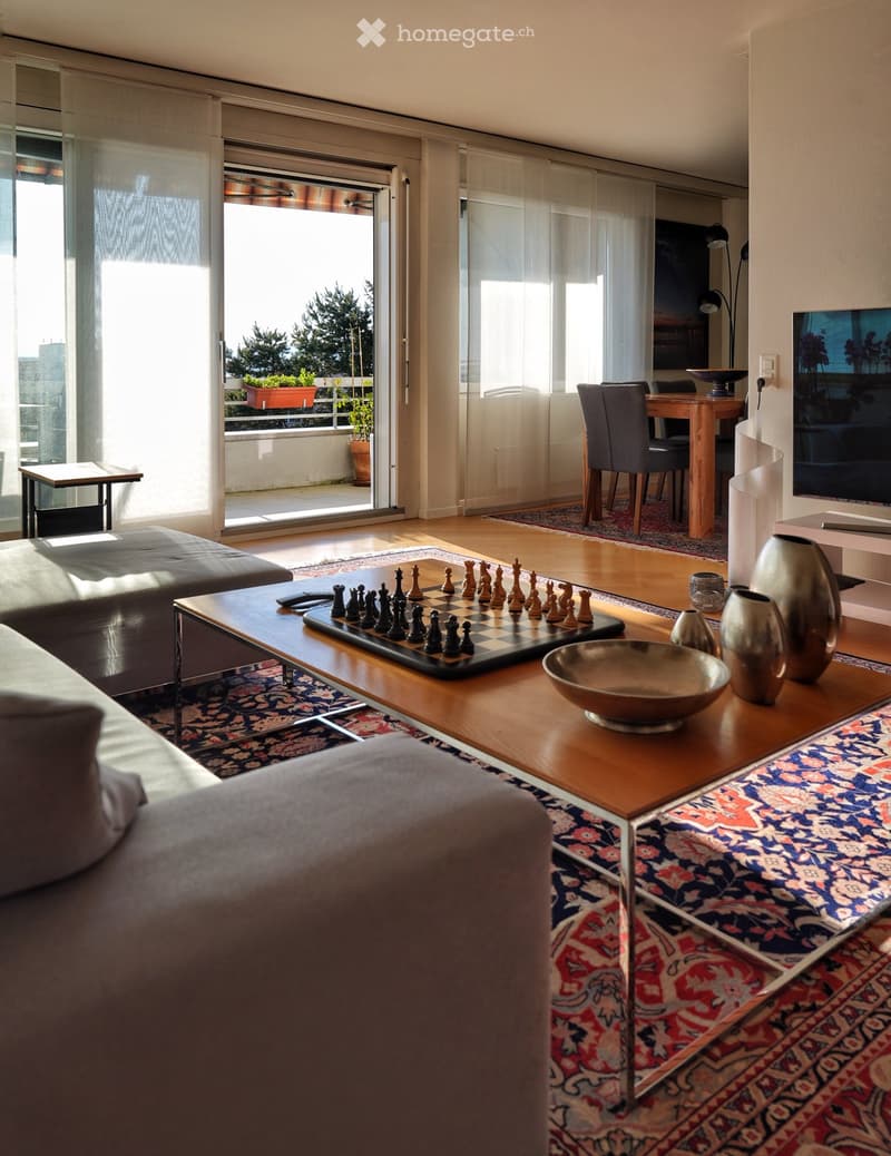 Modern living room with large windows and a view, featuring a grey sofa, wooden coffee table with chessboard, decorative vases, and a dining area with chairs and a table.