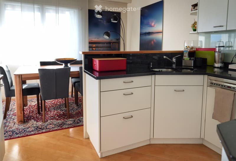 Modern kitchen with white cabinets, black countertops, a sink, and a small dining area with a wooden table and chairs. The space features a blender, a bowl, and a bottle on the counter.