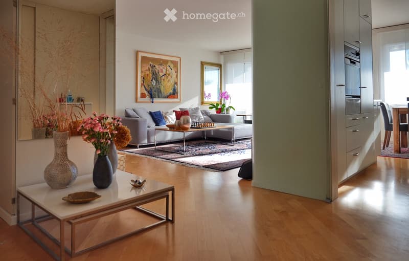 Modern living room with light wooden flooring, a grey sectional sofa, a white coffee table, decorative vases, and a dining area visible in the background.