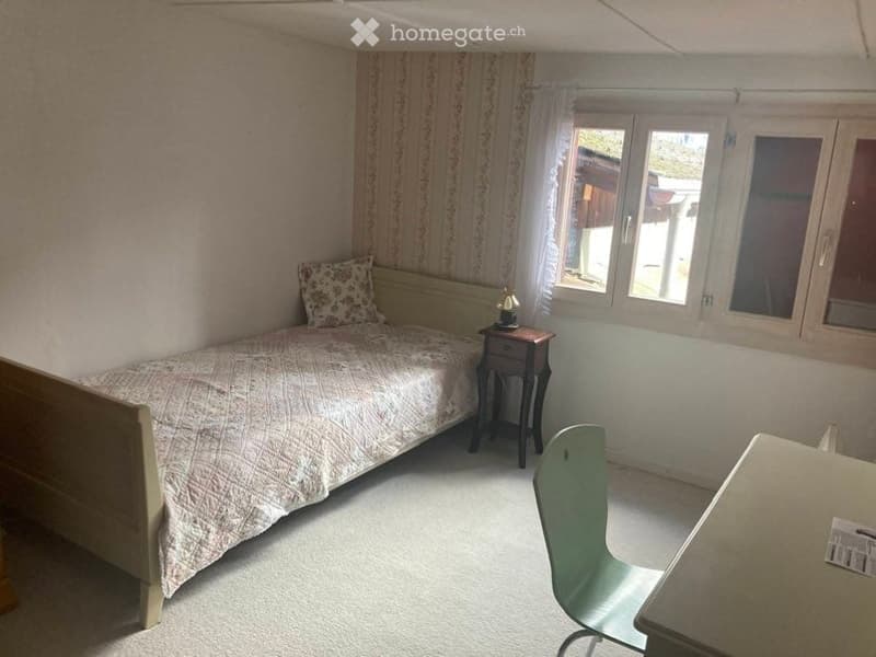 Cozy bedroom with a single bed, nightstand, chair, and small table. Light-colored walls and carpeted floor.