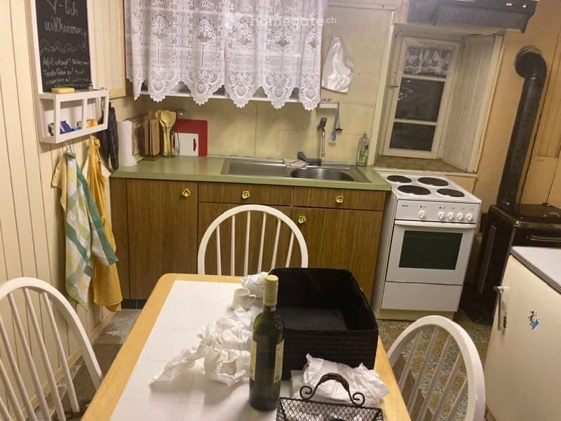 A small kitchen with wooden cabinets, a sink, a stove, and a dining table with chairs. There is a bottle and some cups on the table, along with a black tray and a small basket.
