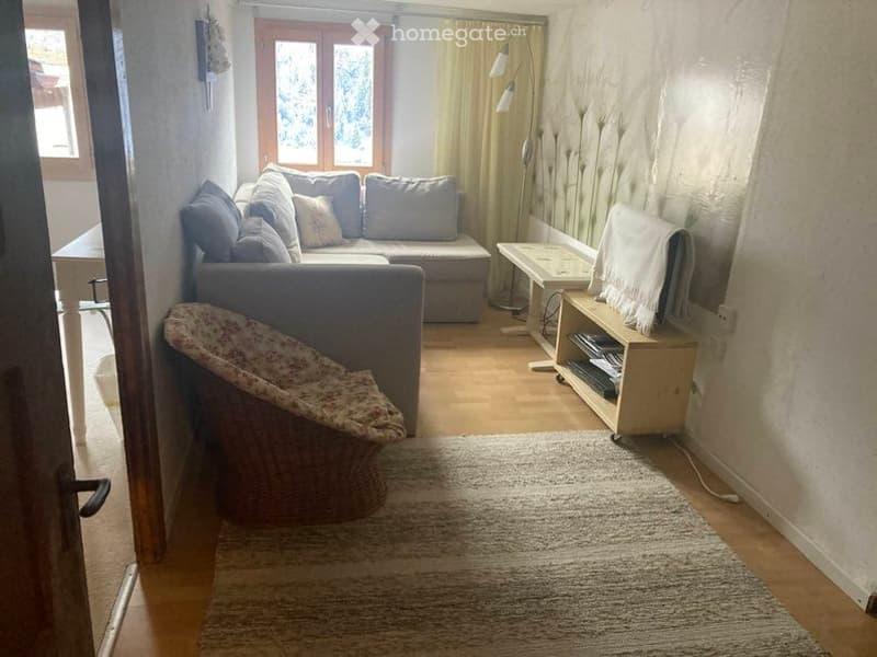 Cozy living room with a grey sectional sofa, coffee table, wicker chair, floor lamp, and TV stand. Light-colored walls and wooden flooring.