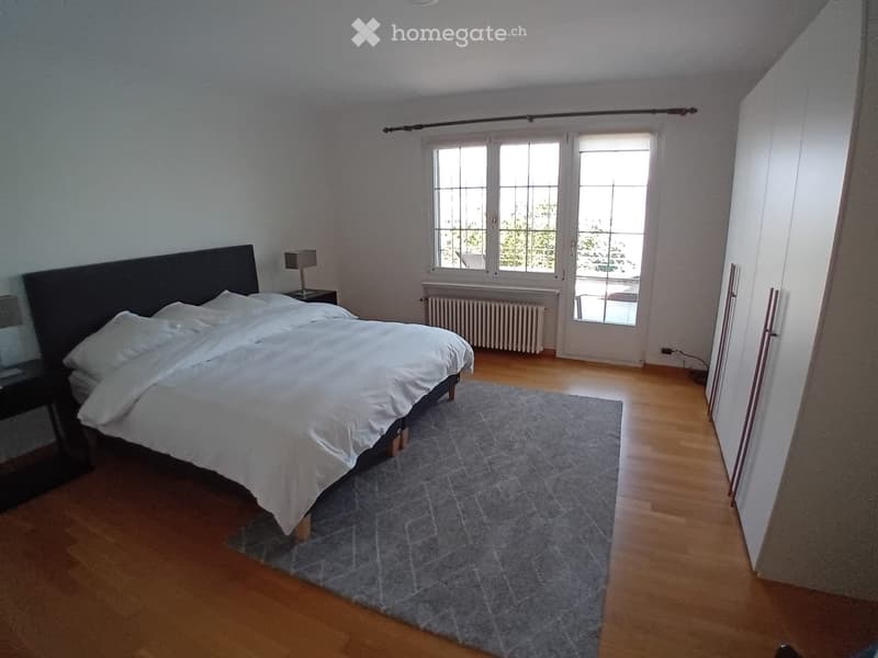 Bright bedroom with wooden flooring, large windows, a neatly made bed with white linens, a nightstand with a lamp, and a grey rug.