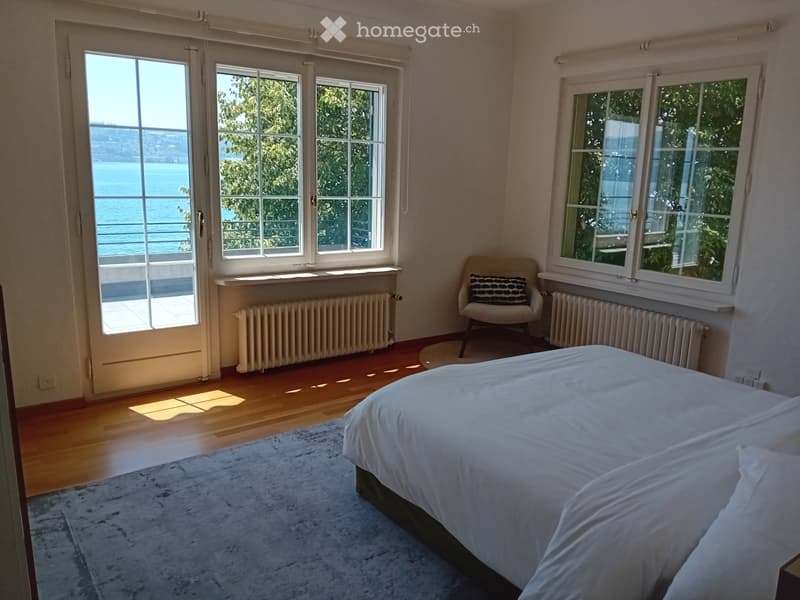 Bright bedroom with large windows overlooking a scenic view, featuring a bed, a chair, and a rug.