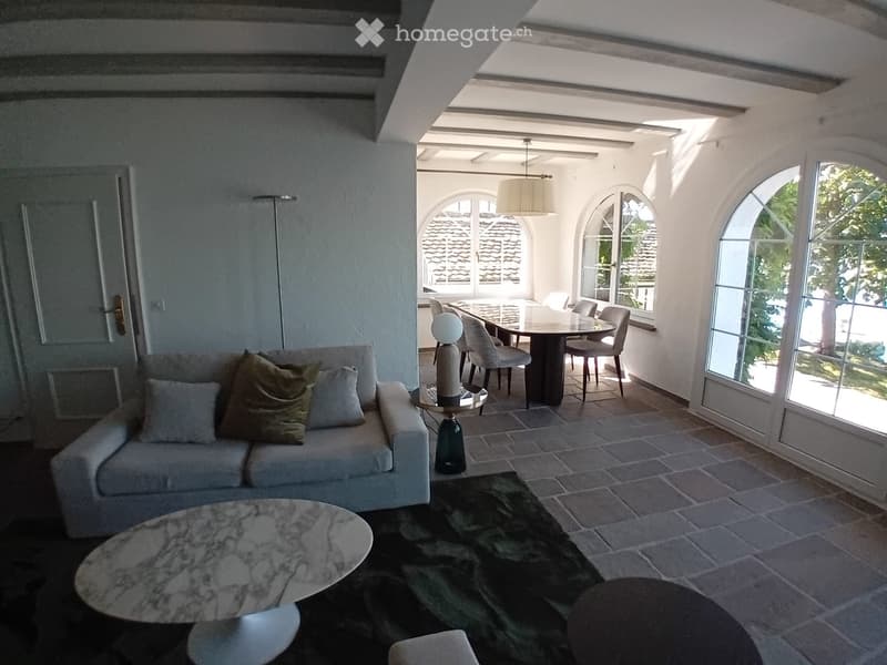 Bright living room with a sofa, coffee table, dining table, and chairs. Large arched windows and a hanging lamp provide natural light.