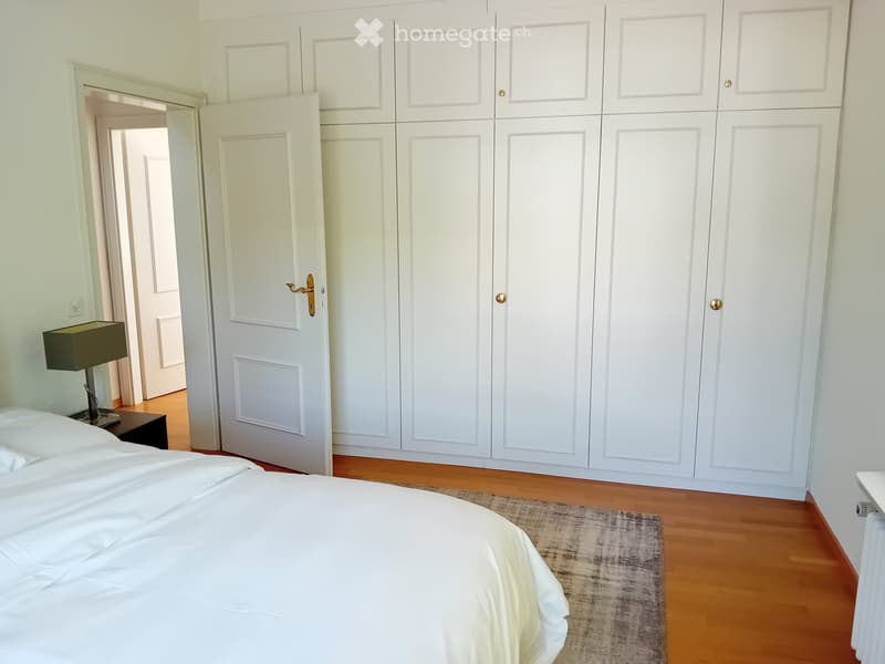 A well-lit bedroom with a large bed, nightstand, lamp, and a large white wardrobe with multiple doors.