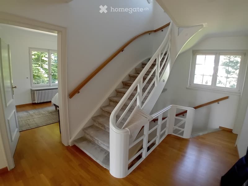 Spacious hallway with wooden flooring, a curved staircase with white railings, and large windows letting in natural light. The hallway features a radiator and a glimpse of a room with a bed and rug visible in the background.