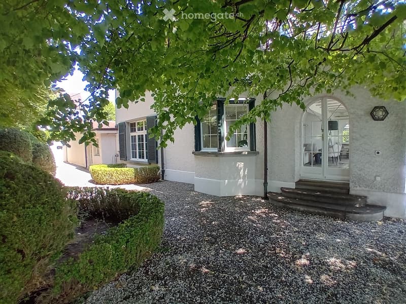 Garden area with gravel pathway, trimmed bushes, and a house with white walls and black shutters visible through trees.