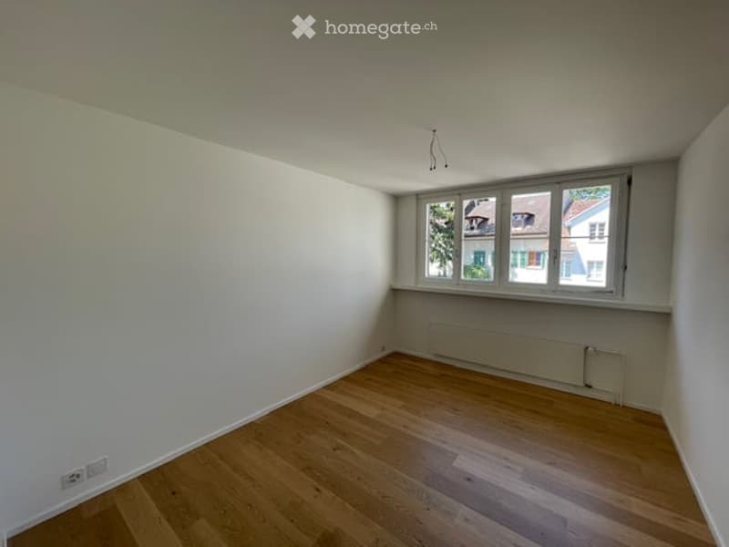 Empty room with wooden flooring, white walls, and large windows showing a view of neighboring houses.